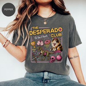 May include: A charcoal grey t-shirt featuring a graphic print with the text THE DESPERADO CLUB, So fun it hurts, Mongo no!, and Goddammit Donut. The design includes a gold trophy, patterned shorts, a donut, a cat, a dinosaur, a sword, and a skull.