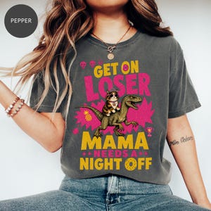 May include: A charcoal gray t-shirt featuring a graphic of a cat wearing sunglasses riding a green dinosaur. The text reads GET ON LOSER MAMA NEEDS A NIGHT OFF. The design includes pink and yellow accents, a pineapple, a trophy, and small skulls.