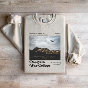 May include: Cream-colored sweatshirt with a graphic featuring a mountain landscape, a flying dragon, and the text "Basgiath War College." Additional text includes "Threshing Day, October 1st" and "Only Dragon Riders Will Survive."