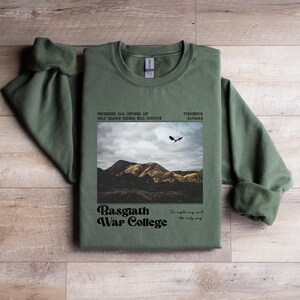 May include: An olive green sweatshirt with the text "THURSHING DAY, OCTOBER 1ST ONLY DRAGON RIDERS WILL SURVIVE" and "Basgiath War College". The design includes a landscape image with a flying dragon.