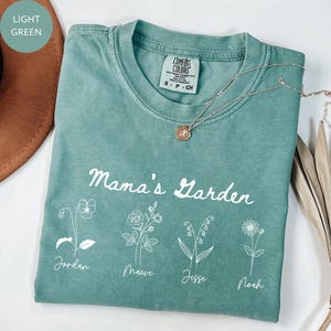 May include: A light green t-shirt with the words "Mama's Garden" and floral illustrations. The names Jordan, Maeve, Jesse, and Noah are printed below the flowers. A silver necklace with a pendant is draped across the shirt.