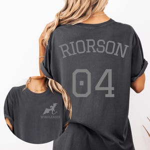 May include: Dark gray t-shirt with the name "RIORSON" and the number "04" printed on the back in gray. A smaller image of a dragon and the word "WINGLEADER" are on the front. The shirt has short sleeves.