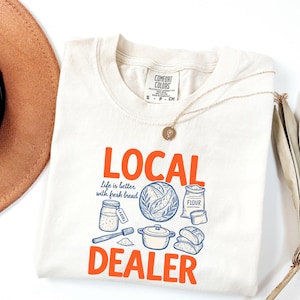 May include: Cream-colored t-shirt with the words "LOCAL DEALER" in orange, and a graphic of bread-making tools and ingredients in blue. The shirt also has the text "life is better with fresh bread."