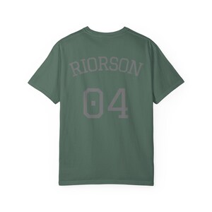 May include: A sage green short-sleeve t-shirt featuring the text RIORSON arched above the number 04 in a dark grey block font. The garment has a crew neckline and a standard fit, displaying a clean design on the back of the shirt.