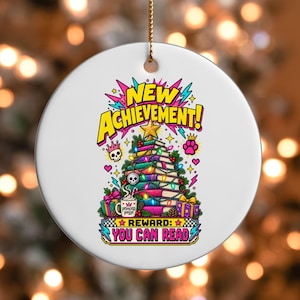 May include: A round white ceramic ornament featuring a colourful graphic of a stack of books shaped like a Christmas tree, topped with a gold star. Text reads NEW ACHIEVEMENT! and REWARD: YOU CAN READ, with illustrations of a skull, gifts, and a mug.