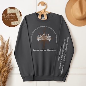 May include: A dark grey crewneck sweatshirt featuring a white graphic of a crown with the text "DEVOURER OF CROWNS, RAVAGER OF REALMS, HERALD OF VENGEANCE" and "DAUGHTER OF THE FORGOTTEN". The sleeve includes the text "I WANT TO KNEEL AT YOUR FEET, SOAKED IN THE BLOOD OF YOUR ENEMIES... -LUTHER CORBOIS".