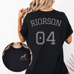 May include: A black t-shirt featuring the text RIORSON and the number 04 in grey on the back. The front chest area displays a small grey graphic of a dragon silhouette above the text WINGLEADER, shown in a circular inset on the lower left.