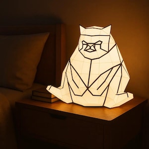 Pode incluir: Uma lâmpada geométrica em forma de gato com uma sombra branca e contornos pretos. A lâmpada está em uma mesa de cabeceira de madeira. A lâmpada está acesa, lançando um brilho quente na sala. É um item decorativo único.