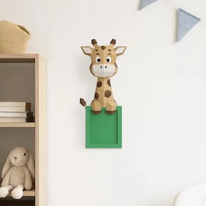 May include: A papercraft giraffe with brown spots and a green frame. The giraffe is mounted on a white wall, with a shelf and stuffed bunny in the background. Blue triangle flags hang from the ceiling.