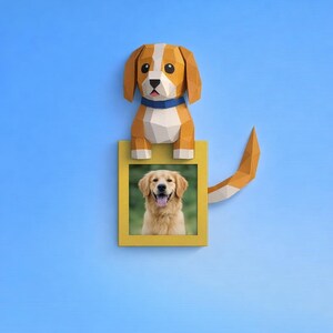May include: A papercraft dog in shades of brown and white perches above a yellow picture frame. The frame contains a photo of a golden retriever. The dog wears a blue collar and has a tail. The background is a light blue.