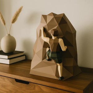 Peut inclure: Une sculpture géométrique marron représentant un grimpeur escaladant une montagne. Le grimpeur porte un haut marron et un short vert. La montagne est de couleur marron clair. La sculpture est sur une surface en bois.
