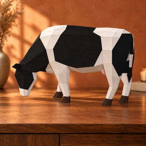 Op de afbeelding: Een low-poly papieren sculptuur van een koe met een geometrisch ontwerp met zwarte en witte vlekken. Het figuur staat in een grazende houding met donkerbruine hoeven en een kleine roze uier zichtbaar aan de onderkant van het lichaam.