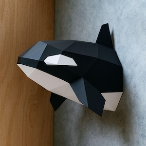 Op de afbeelding: Een geometrische orka-walvishoofd van papier, gemonteerd aan een muur. De sculptuur is voornamelijk zwart en wit, met scherpe hoeken en een moderne esthetiek. Het oog van de orka is een witte polygoonvorm.