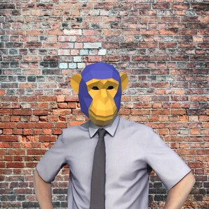 May include: A person wearing a geometric monkey mask with blue and yellow accents, a light gray shirt, and a dark gray tie, standing in front of a brick wall.