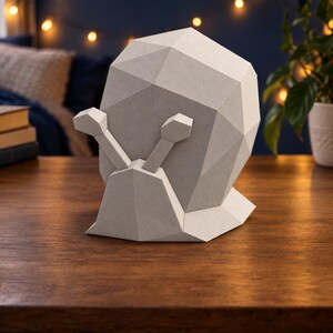 Op de afbeelding: Een lichtgrijze papieren sculptuur van een slak met een geometrisch, gefacetteerd ontwerp. De slak heeft een grote, ronde schelp die is opgebouwd uit meerdere platte driehoekige vlakken en twee uitstekende oogstelen op de kop.
