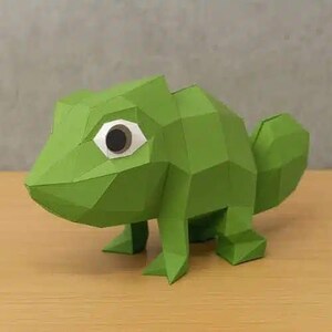 May include: A green paper chameleon sculpture with a geometric, faceted design. The chameleon has a large eye with a black pupil and white highlights. The sculpture is standing on four legs and has a curled tail. The background is a neutral grey.