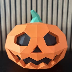 Peut inclure: Une citrouille décorative orange en low-poly avec une surface géométrique à facettes, une tige sarcelle et des découpes sombres pour les yeux, le nez et la bouche dentelée. La structure est composée de plans plats et angulaires.