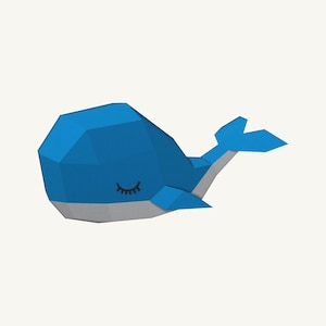 Może przedstawiać: Figurka wieloryba z papieru w stylu low-poly z jasnoniebieskim grzbietem i jasnoszarym brzuchem. Wieloryb ma geometryczny, fasetowany kształt z zamkniętym okiem przedstawionym za pomocą czarnych, zakrzywionych linii i rzęs na boku.