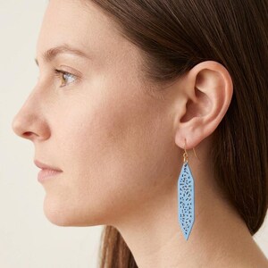 May include: A close-up of a person wearing a light blue, leaf-shaped earring. The earring has a delicate, openwork design and a gold-colored hook. The earring is hanging from the ear.