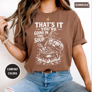 May include: Espresso-colored t-shirt with the text "THAT'S IT YOU'RE GOING IN THE SOUP" and a graphic of a Bigfoot-like creature stirring a cauldron. The shirt is a comfort color and has rolled-up sleeves.