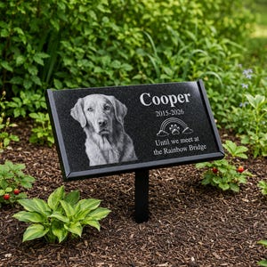 May include: A black granite rectangular pet memorial plaque on a metal stake. It features an engraved portrait of a dog, the name Cooper, the years 2015-2026, a rainbow icon with a paw print, and the text Until we meet at the Rainbow Bridge.