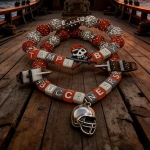 May include: Two beaded bracelets featuring red, white, and silver rhinestones, orange and white letter beads spelling TAMPA BAY BUCCANEERS, a skull and crossbones charm, two small ship charms, and a silver-toned football helmet pendant.