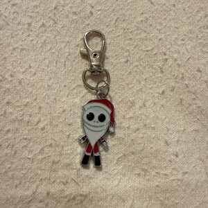 Peut inclure: Un porte-cl&eacute;s en m&eacute;tal argent&eacute; avec une breloque repr&eacute;sentant un personnage v&ecirc;tu d'un costume de P&egrave;re No&euml;l rouge et blanc. Le personnage a un visage blanc avec des yeux noirs circulaires et une bouche cousue, portant un bonnet rouge.