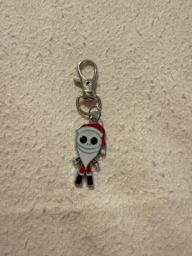 Peut inclure: Un porte-cl&eacute;s en m&eacute;tal argent&eacute; avec une breloque repr&eacute;sentant un personnage v&ecirc;tu d'un costume de P&egrave;re No&euml;l rouge et blanc. Le personnage a un visage blanc avec des yeux noirs circulaires et une bouche cousue, portant un bonnet rouge.