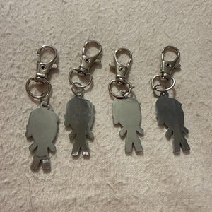 Peut inclure: Quatre porte-cl&eacute;s en m&eacute;tal argent&eacute;, chacun pr&eacute;sentant une petite silhouette de personnage stylis&eacute;e attach&eacute;e &agrave; un fermoir mousqueton. Les figurines ont une surface lisse et r&eacute;fl&eacute;chissante et sont dispos&eacute;es en rang&eacute;e, avec une finition m&eacute;tallique.