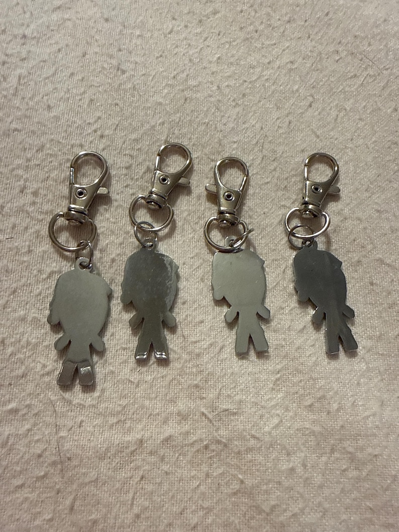 Peut inclure: Quatre porte-cl&eacute;s en m&eacute;tal argent&eacute;, chacun pr&eacute;sentant une petite silhouette de personnage stylis&eacute;e attach&eacute;e &agrave; un fermoir mousqueton. Les figurines ont une surface lisse et r&eacute;fl&eacute;chissante et sont dispos&eacute;es en rang&eacute;e, avec une finition m&eacute;tallique.