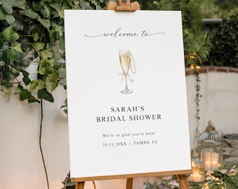 Puede incluir: Un letrero de bienvenida rectangular blanco para una despedida de soltera sobre un caballete de madera. El letrero presenta el texto "welcome to", "SARAH'S BRIDAL SHOWER", "We're so glad you're here!" y "10.31.20XX | TAMPA, FL" con una ilustración.