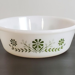 May include: A white milk glass bowl featuring a green floral pattern around the exterior. The bowl has a round shape with two small integrated handles on opposite sides. The design consists of stylized flowers and leafy vines in dark green.
