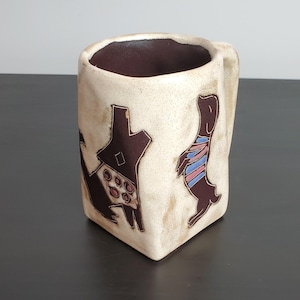May include: A square-shaped ceramic mug with a cream-colored, speckled exterior and a dark brown interior. The mug features two dark brown animal silhouettes: one wearing a patterned garment with red circles and one wearing a striped shirt.
