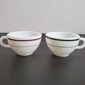 May include: Two white milk glass cups with rounded handles. One cup features a single horizontal maroon stripe around the center, while the other cup features a single horizontal dark green stripe around the center. Both cups have a smooth finish.