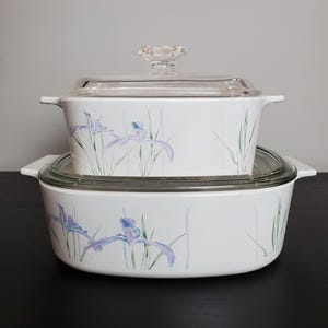 May include: Two white rectangular casserole dishes with integrated side handles and clear glass lids. The exterior of each dish features a delicate botanical pattern of light purple iris flowers and green stems against a clean white background.