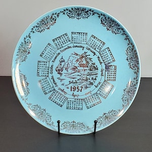 May include: A vintage 1957 calendar plate with a light blue background and gold accents. The plate features a calendar around the edge, a windmill scene, and decorative floral patterns. The plate is displayed on a black stand.