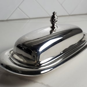 May include: A polished silver-tone metal butter dish with an oval base and a matching domed lid. The lid features a decorative finial shaped like a small pineapple. The surface of the metal is highly reflective, showing smooth, curved lines.