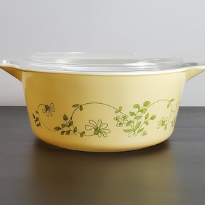 May include: A round, pale yellow casserole dish with integrated side handles and a clear glass lid. The exterior features a repeating green botanical pattern of delicate flowers, leaves, and vines encircling the center of the bowl.