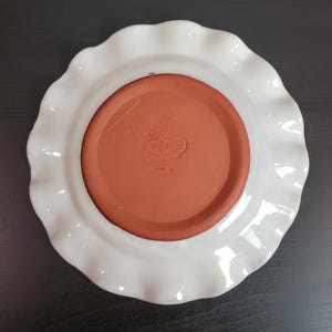 May include: A round ceramic plate featuring a white glazed ruffled rim and a contrasting unglazed terracotta center. The center base displays an embossed maker's mark with the year 1983 and a small number 8 printed on the white outer edge.