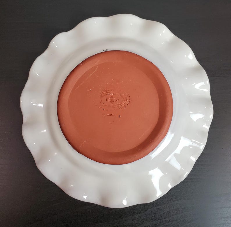 May include: A round ceramic plate featuring a white glazed ruffled rim and a contrasting unglazed terracotta center. The center base displays an embossed maker's mark with the year 1983 and a small number 8 printed on the white outer edge.