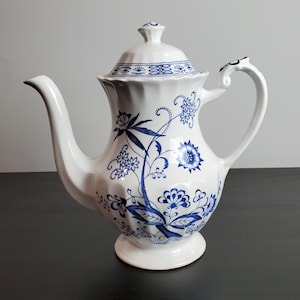 May include: A white porcelain coffee pot featuring a blue floral pattern, a scalloped body, a curved handle, and a matching lid. The design includes intricate blue botanical illustrations of flowers and leaves, with a flared base and spout.