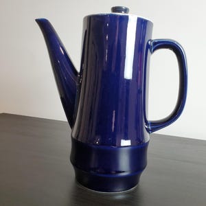 May include: A tall, cylindrical ceramic coffee pot with a glossy, deep cobalt blue glaze. The vessel features a long, angled spout, a curved handle on the side, and a matching lid with a small knob. The base has a slightly wider, tiered design.