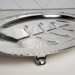 Op de afbeelding: Een ovale zilverkleurige serveerschaal met een gepolijst, reflecterend oppervlak. Het midden is voorzien van een verzonken, in reliëf aangebracht bladpatroon. De rand heeft een decoratieve kralenafwerking en de schaal rust op vier korte, gebogen pootjes.