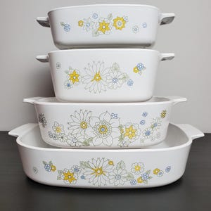 May include: A set of four white rectangular baking dishes stacked vertically, each featuring integrated side handles and a decorative floral pattern in yellow, blue, and green line art. The dishes vary in size, decreasing from bottom to top.
