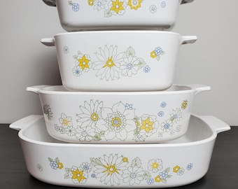 Op de afbeelding: Een set van vier witte rechthoekige ovenschaaltjes verticaal gestapeld, elk voorzien van geïntegreerde handgrepen aan de zijkant en een decoratief bloemenpatroon in gele, blauwe en groene lijntekeningen. De schalen variëren in grootte van onder naar boven.