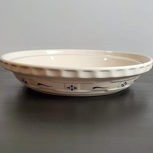 May include: A round, cream-colored ceramic pie dish with a fluted rim. The exterior features a repeating pattern of small, dark blue floral motifs and horizontal lines encircling the side of the dish, providing a decorative accent to the piece.