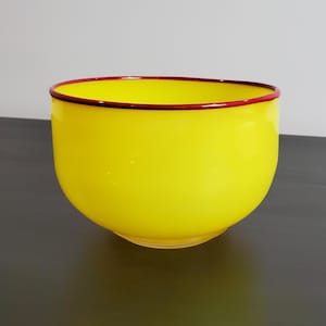 May include: A bright yellow glass bowl with a smooth, rounded shape and a distinct, thin dark red rim encircling the top edge. The bowl features a translucent quality and a polished finish, showcasing a simple, curved design without any handles.