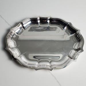 Könnte beinhalten: Ein poliertes silberfarbenes Serviertablett mit einer reflektierenden, spiegelartigen Oberfläche. Das Tablett hat eine ovale Form mit einem gewellten, dekorativen Rand und einer glatten, flachen Mitte. Das Metallfinish erzeugt Lichtreflexe.