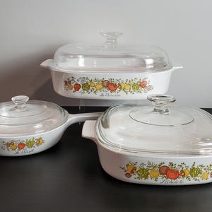 May include: A set of three white ceramic cookware pieces with clear glass lids. Each piece features a colorful vegetable pattern and French text labels including Le Persil, Le Romarin, and L'Echalote. The set includes a skillet and two casseroles.