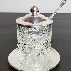 May include: A clear glass sugar bowl with a silver lid and a small spoon. The bowl has a diamond pattern and sits on a silver plate. The spoon is resting on the lid.